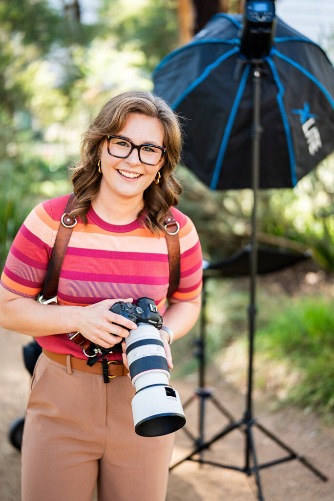 Photo of Alexandra Orme of Alexandra Orme Photography smiling at the camera and holding her camera with light in the background